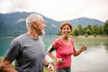 Senior pensioner couple with smartphone running by lake in nature.