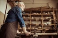 Senior potter in apron standing and leaning on table against shelves with pottery goods at workshop Royalty Free Stock Photo