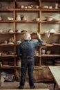 Senior potter standing near shelves with pottery goods and searching something at workshop