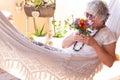 Senior pretty woman with gray hair lying on a white hammock and sniffs a bouquet of flowers. Relaxing moment outdoors on the