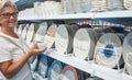 Senior pretty woman with gray hair. Standing and choosing a plate or a bowl for the table. In the background a large selection of