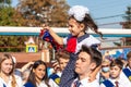 Senior pupil carries first-former girl ringing with golden bell against schoolchildren. Tradition of beginning of elementary