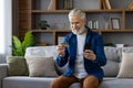 Senior smiling gray-haired man sitting on sofa at home and using phone and credit card Royalty Free Stock Photo