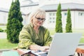 Senior beautiful business woman sitting on a chair and making notes in laptop with a notebook, with a phone in the yard Royalty Free Stock Photo