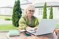 Senior beautiful business woman sitting on a chair and making notes in laptop with a notebook, with a phone in the yard Royalty Free Stock Photo