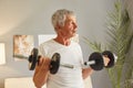 Senior sport grey-haired mature man using barbels foe training biceps and triceps having home workout wearing casual white T-shirt Royalty Free Stock Photo