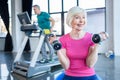 senior sportswoman sitting on fitness ball with dumbbells sportsman on treadmill behind in senior fitness class