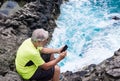 Senior sporty man in outdoor excursion at sea sitting on the cliff using mobile phone. White hair and beard