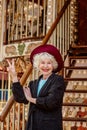 Senior Stylish Woman In Dark Grey Coat, Hat And With Grey Hair Standing By The Carousel Smiling And Enjoying Life.