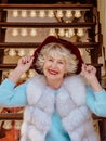 Senior stylish woman in fur coat and with grey hair sitting on carousel holding purple hat.