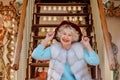 Senior stylish woman in fur coat and with grey hair sitting on carousel holding purple hat.