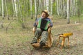 Senior wearing straw hat singing and playing garmonika while sitting lonely in wicker rocking chair in birch forest