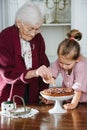 Senior woman along with her granddaughter pouring powdered sugar on tasty pie