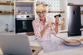 Senior woman in apron and with eyeglasses standing in kitchen, using laptop and preparing healthy meal Royalty Free Stock Photo