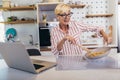 Senior woman in apron and with eyeglasses standing in kitchen, using laptop and preparing healthy meal Royalty Free Stock Photo