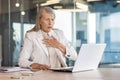 Senior woman in business suit sitting at desk in office in front of laptop and holding hand to chest, having panic Royalty Free Stock Photo