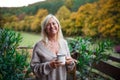 Senior woman with coffee standing outdoors on terrace, looking at camera.