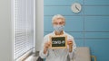 Senior woman doctor with disposable mask holds blackboard with word Covid 19 sitting in contemporary hospital office