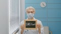 Senior woman doctor with disposable mask holds blackboard with word Covid 19 sitting in contemporary hospital office