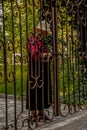 Senior woman in dress and hat locks wrought iron gate at property entrance with chain and padlock - lush vegetation and