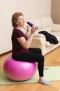 Senior woman exercising on fitness ball, drinking from reusable bottle at home with her schnauzer dog. Active elderly.