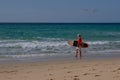 Senior woman with gray hair and a orange top carry a surfboard on the beach