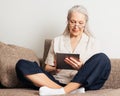 Senior woman with grey hair wearing glasses sitting with crossed legs on a couch using a digital tablet Royalty Free Stock Photo