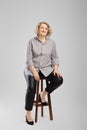 Senior woman in grey shirt and faux leather trousers sits on tall wooden chair in studio Royalty Free Stock Photo