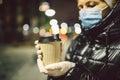 Senior woman holding coffee to go, wearing protective medical mask and white latex gloves, walk outdoors at night, observing Royalty Free Stock Photo