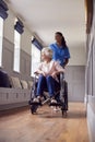 Senior Woman At Home Being Pushed In Wheelchair By Female Care Worker In Uniform