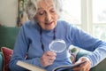 Senior Woman At Home Reading Book Using Magnifying Glass Royalty Free Stock Photo