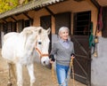 Senior woman horse breeder leading white horse through horse barn Royalty Free Stock Photo