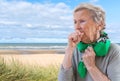 Senior woman l has bad cough, uses tissue, wears scarf on neck, feels unwell and unhealthy seaside background