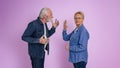 Senior woman looking at camera and showing stop gesture to furious husband arguing with her while standing isolated on purple