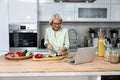 Senior woman looking for cooking recipes online on her laptop. Pensioner female cook vegetables as vegan food standing in the