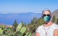 Senior woman in medical mask traveling in mountain landscape in Tenerife, horizon over the sea - new normal life concept in summer