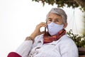 Senior woman outdoors using her cell phone wearing a home made face mask during the coronavirus quarantine de-escalation