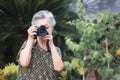 Senior woman with short gray hair shooting photos by a digital camera while standing outdoors. Space for text. Royalty Free Stock Photo