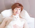 A senior woman with short red hair and a cute white cat with different eyes cozy sitting on a bed