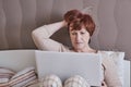 Senior woman with short red hairis sitting on the bed with a laptop, carefully looking at the monitor