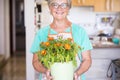 Senior woman showing a jar with a plant with a lot of flowers inside - 60s, retired and mature woman at home - gardener lifstyle