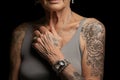 A senior woman in a sleeveless top reveals her heavily tattooed arms and shoulders, with a delicate silver chain around her neck