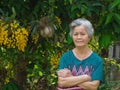 Senior woman smiling, arms crossed and looking at the camera while standing beside beautiful yellow orchids Dendrobium lindleyi Royalty Free Stock Photo