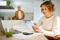 Senior woman using smartphone in front of laptop, working distantly from home when husband cooking at kitchen Royalty Free Stock Photo