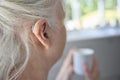 Close Up Of Senior Woman Wearing Behind The Ear Hearing Device Or Aid With Hot Drink At Home