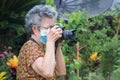 Senior woman wearing a face mask and shooting a photo by the digital camera while standing in a garden. Mask for protecting virus Royalty Free Stock Photo