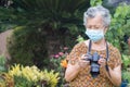 Senior woman wearing a face mask and shooting a photo by the digital camera while standing in a garden. Mask for protecting virus Royalty Free Stock Photo