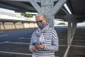 A senior woman wearing flowered medical mask standing under the metal structure of a deserted parking looking at smart phone