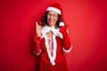 Senior woman wearing Santa Claus costume holding christmas gift over isolated red background pointing and showing with thumb up to Royalty Free Stock Photo