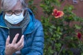 A senior woman wearing a surgical mask due to coronavirus sitting in a blossom garden looking at her smartphone. Old retiree using